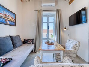 Ein Zimmer mit einer Couch, einem Sessel, einem Tisch mit Platzdeckchen, einem Fernseher und einem Fenster mit Vorhängen.