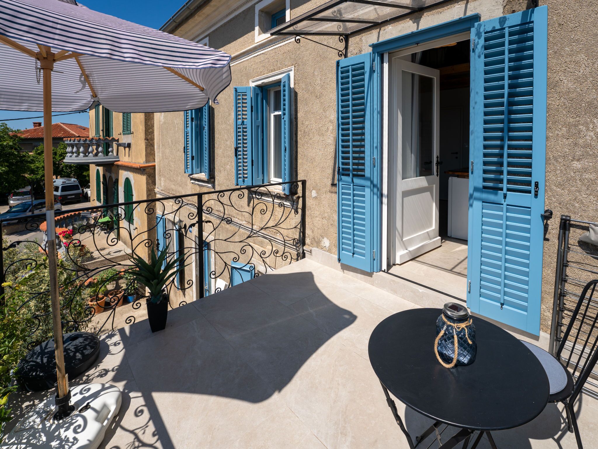 A balcony with a table and chairs, a striped umbrella, and blue shutters on a building.