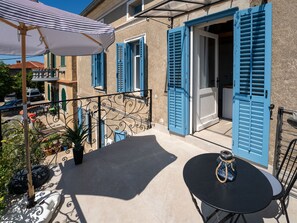 Ein Balkon mit Tisch und Stühlen, einem gestreiften Sonnenschirm und blauen Rollläden an einem Gebäude.