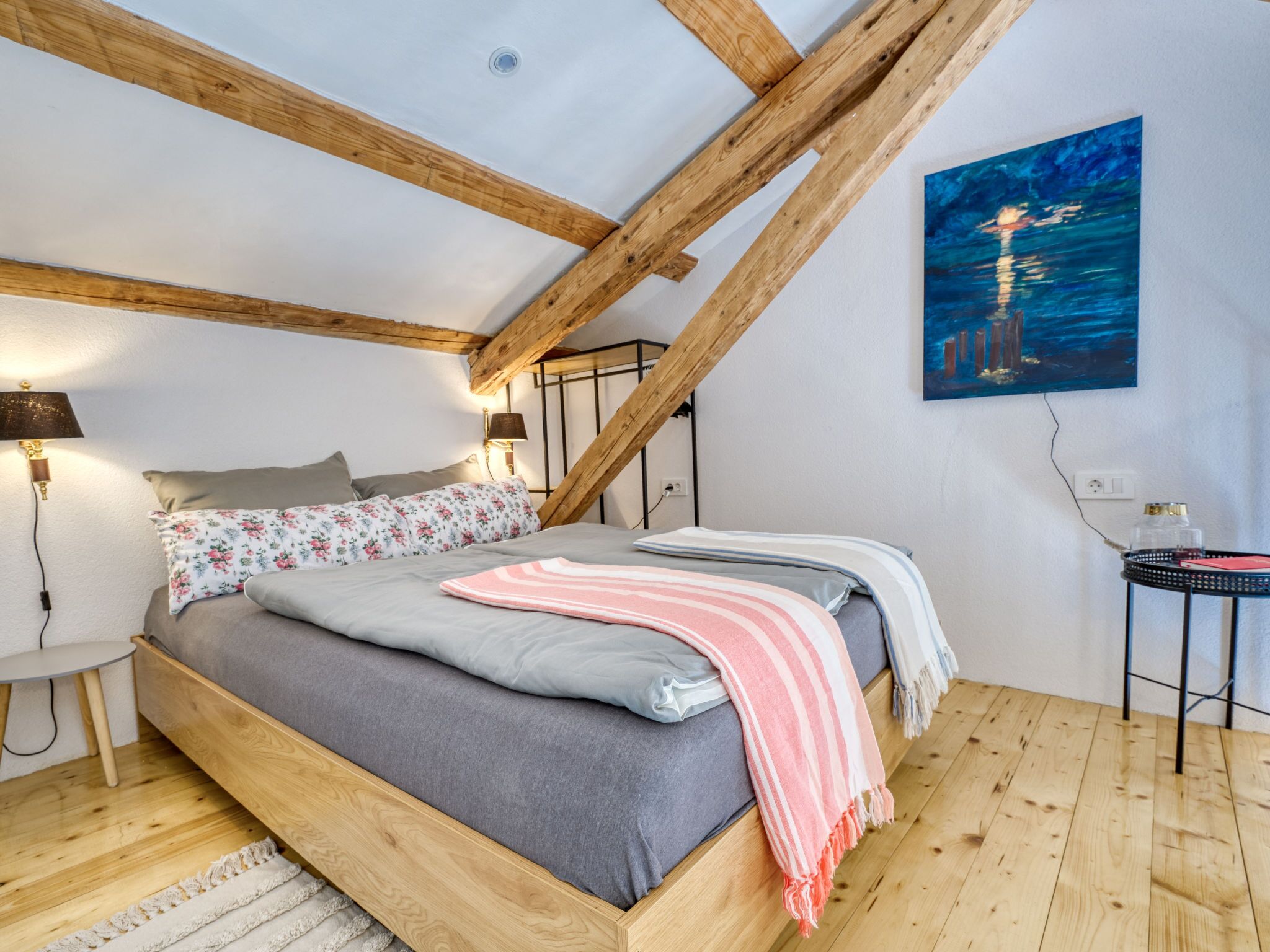 A bedroom with a wooden ceiling, a bed with grey bedding, a pink and white striped blanket, a nightstand with a lamp, and a painting of a cityscape.