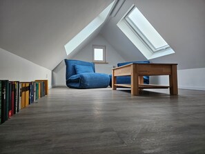 Ein Schlafzimmer im Loft-Stil mit schrägem Dach, einem Oberlicht, einem Holzbett, einem kleinen Tisch und einem Bücherregal.