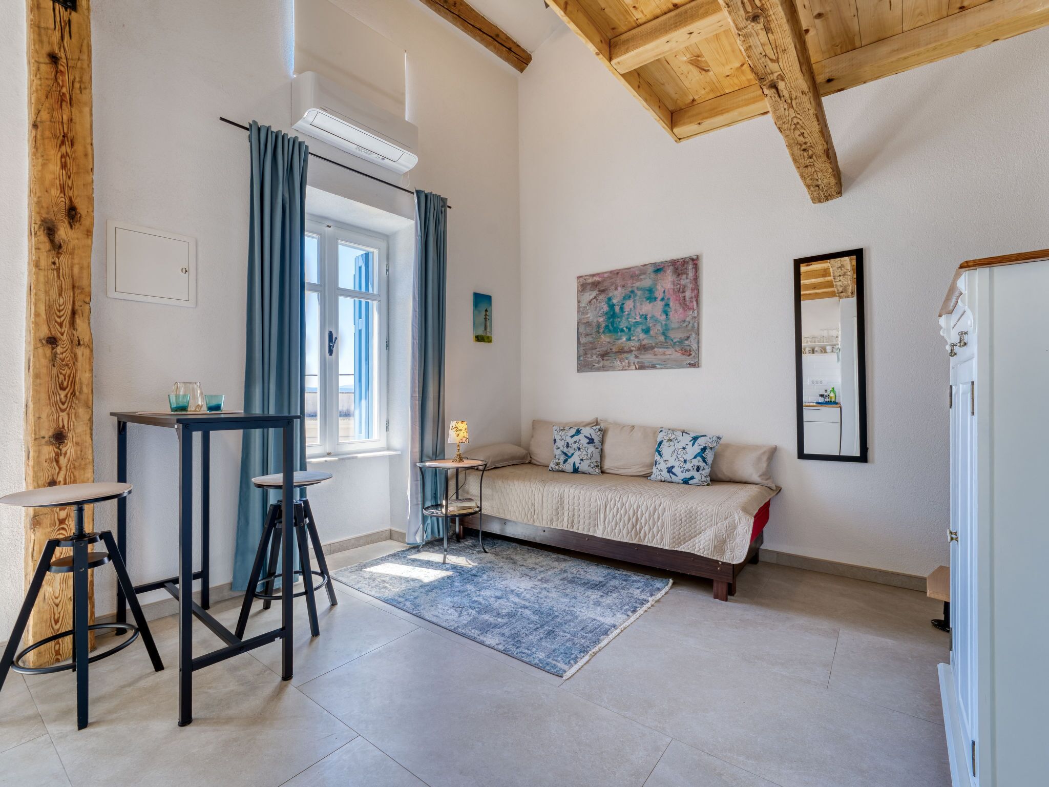 Una habitación con una cama, una mesita, un taburete, una ventana con cortinas, un cuadro y un espejo.