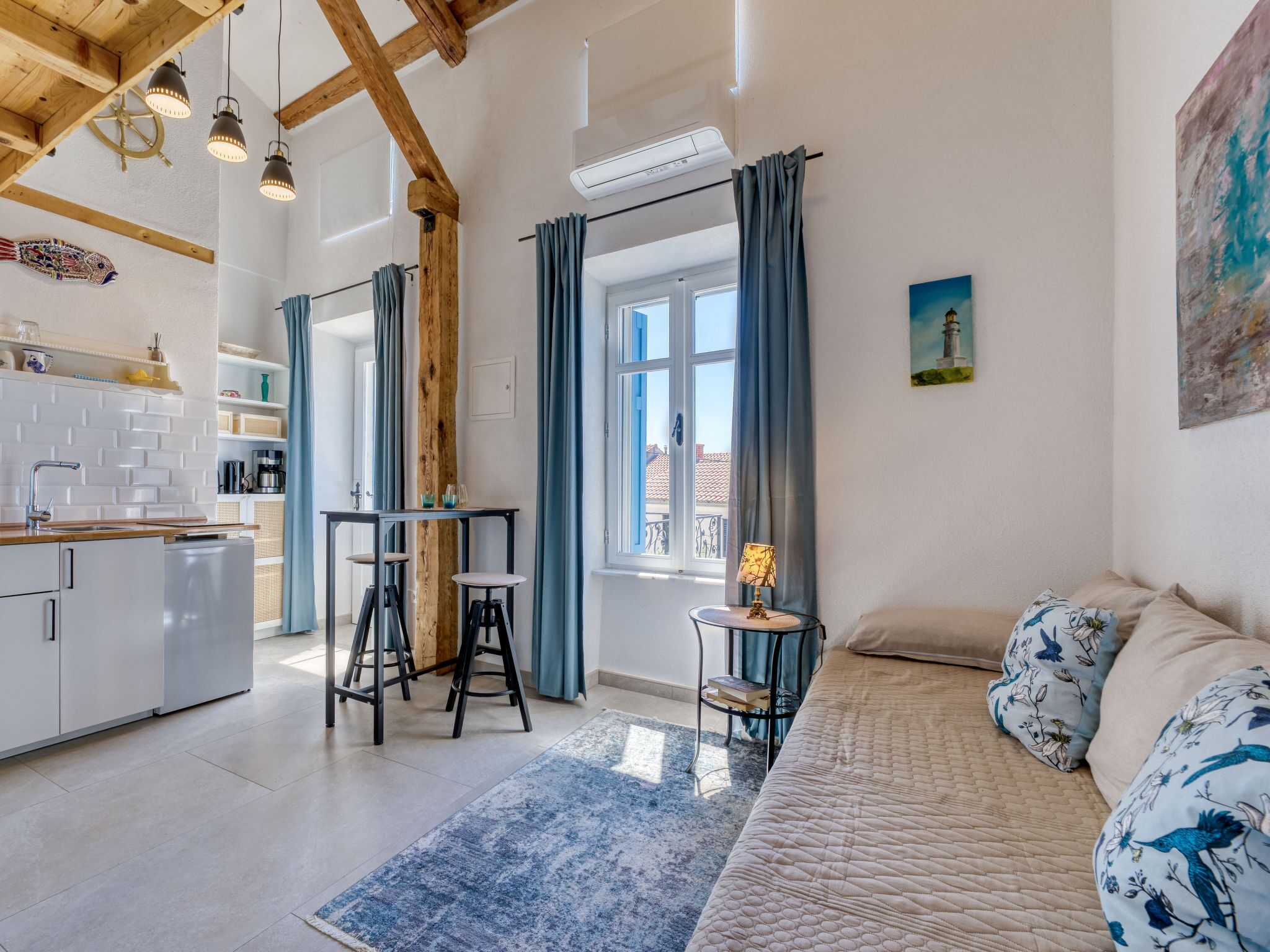 Eine moderne Küche mit weißen Schränken, einer Holzbalkendecke und Pendelleuchten. Ein Essbereich mit Tisch und Stühlen lädt zum Verweilen ein, und ein großes Fenster mit Vorhängen sorgt für eine angenehme Atmosphäre.