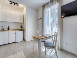 Eine kompakte Küche mit weißen Schränken, einem kleinen Kühlschrank, einem Holztisch mit einem Stuhl und einem großen Fenster mit leichtem Vorhang.