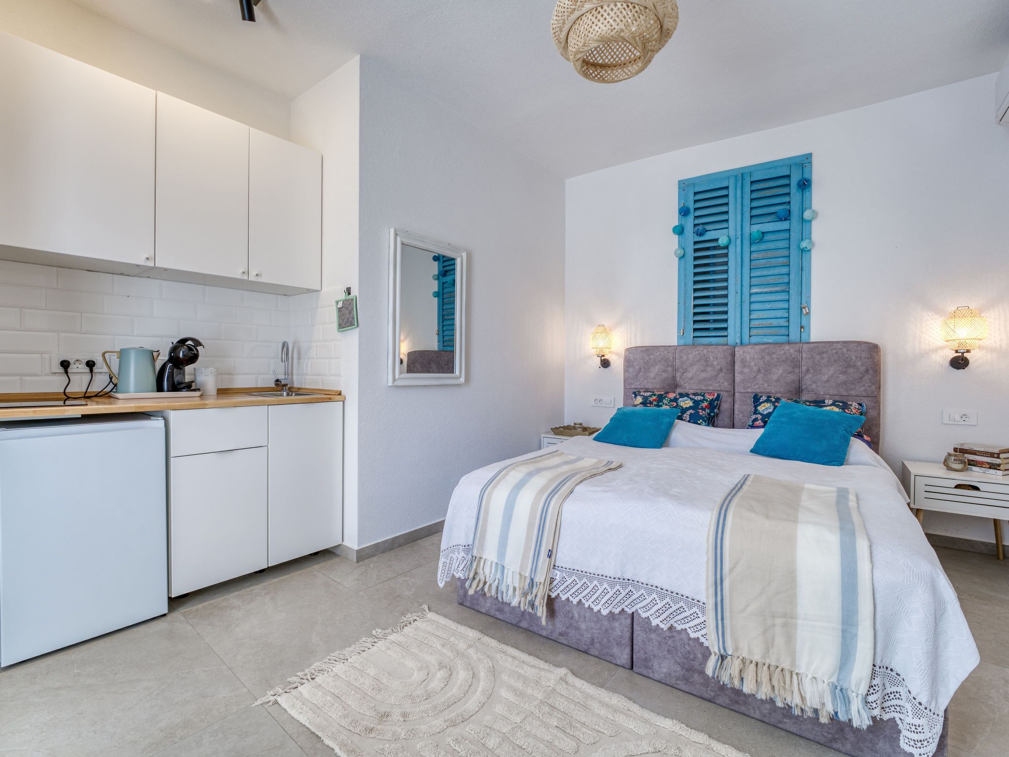 A modern bedroom with a bed, bedside tables, a nightstand, a wall-mounted lamp, and a kitchen area with cabinets and a countertop.