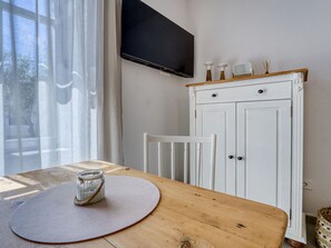 Une pièce avec une table en bois, une armoire blanche, un téléviseur fixé au mur et une fenêtre avec des rideaux.