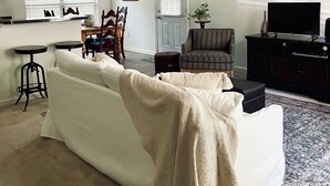 A living room with a white sofa, a ceiling fan, a dining area with a table and chairs, a television, and a patterned rug. - Charming 3-bedroom Monthly Rental Home in Mandeville (Mandeville)
