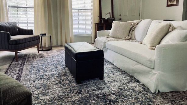 A living room with a sofa, ottoman, and armchair, a patterned rug, and a large window with curtains. - Charming 3-bedroom Monthly Rental Home in Mandeville (Mandeville)