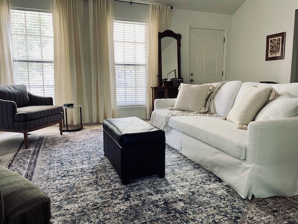 A living room with a sofa, ottoman, and armchair, a patterned rug, and a large window with curtains. - Charming 3-bedroom Monthly Rental Home in Mandeville (Mandeville)