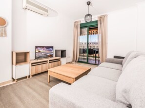 Ein modernes Wohnzimmer mit einer Couch, Fernseher, Couchtisch aus Holz und einer Schiebeglastür, die zu einem Außenbereich führt.