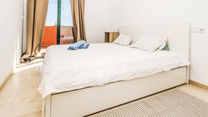 Een slaapkamer met een bed, gordijnen, een nachtkastje en uitzicht op buiten.