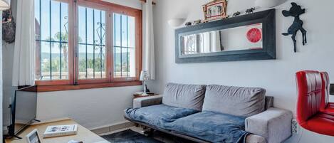A room with a sofa, a red chair, a desk with a lamp, a framed picture, and a window with a view.