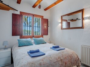 A bedroom with a bed, two pillows, a nightstand, a lamp, a window with shutters, a mirror, and a radiator. - El Refugio de Rembrandt by Interhome (Jávea)