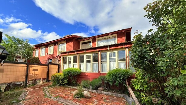 Uma casa vermelha de dois andares com um pátio de tijolos, cercada por vegetação e um muro de madeira.