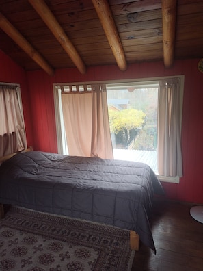 A room with a bed, a window with curtains, a wooden ceiling, and a view of trees outside.