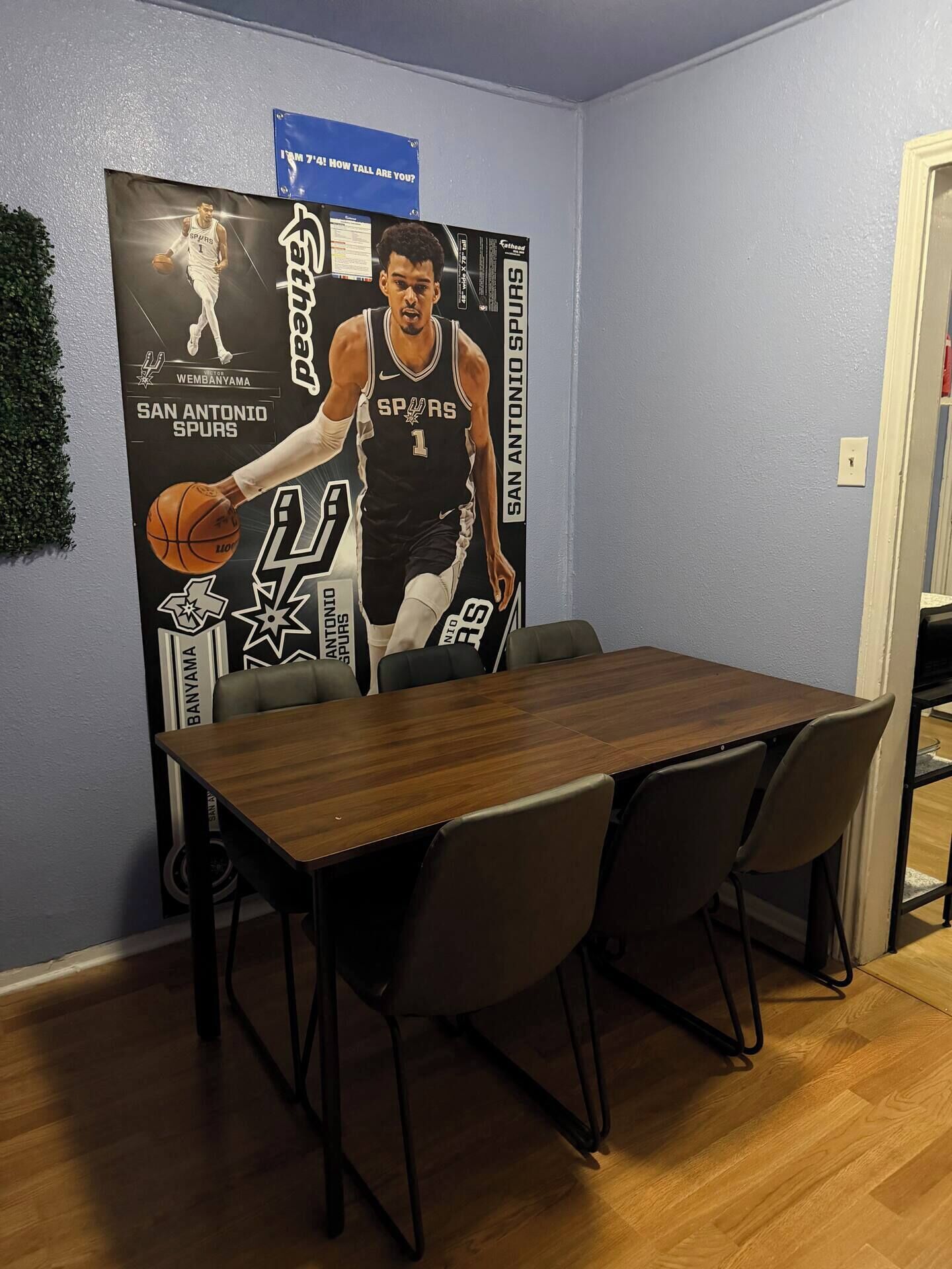 Ein Essbereich mit einem Holztisch und vier Stühlen, einem Poster eines Basketballspielers und einem blauen Schild an der Wand.