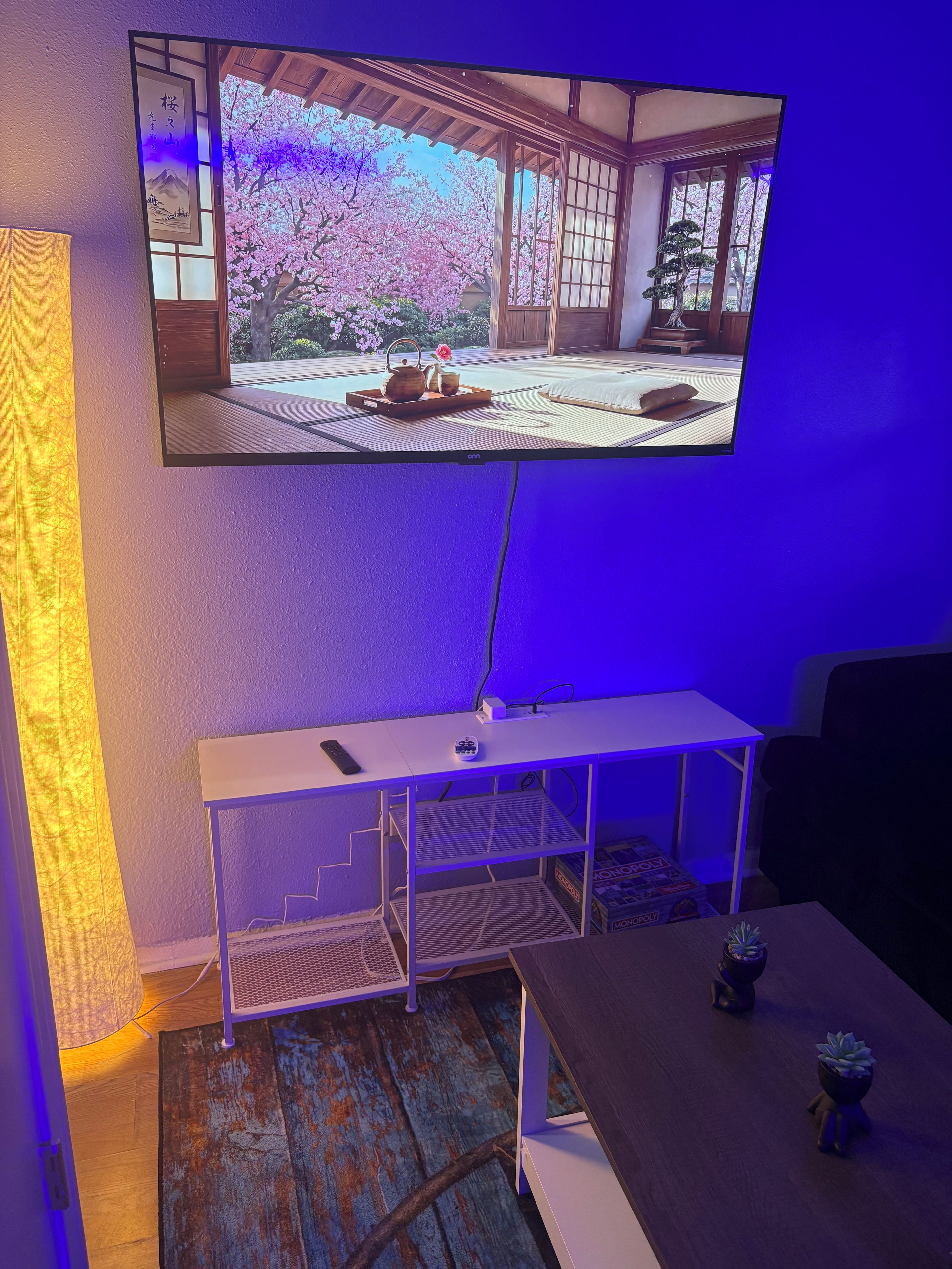Una habitación con una televisión montada en la pared, una mesita consola blanca, un control remoto y una pequeña planta en maceta.