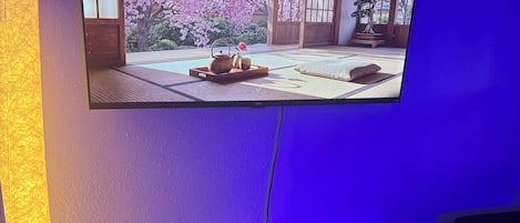 Una habitación con una televisión montada en la pared, una mesita consola blanca, un control remoto y una pequeña planta en maceta.