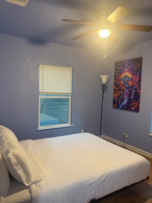 Un dormitorio con una cama, ventilador de techo, ventana con persianas y una lámpara de pared.
