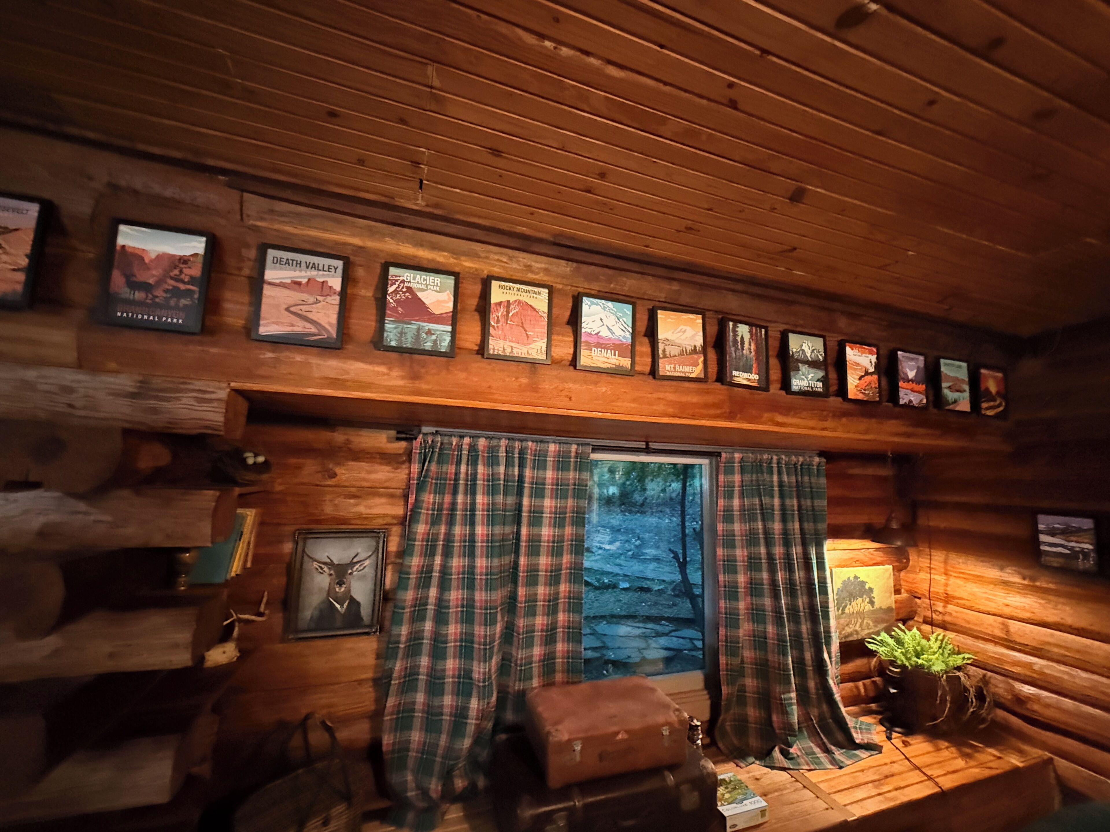 A cabin interior with wooden walls, a window with plaid curtains, a mounted poster of a mountain, and a small table with a plant.