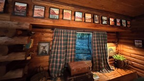 L’intérieur d’une cabane avec des murs en bois, une fenêtre ornée de rideaux à carreaux, une affiche représentant une montagne accrochée au mur et une petite table sur laquelle est posée une plante.