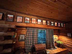 L’intérieur d’une cabane avec des murs en bois, une fenêtre ornée de rideaux à carreaux, une affiche représentant une montagne accrochée au mur et une petite table sur laquelle est posée une plante.