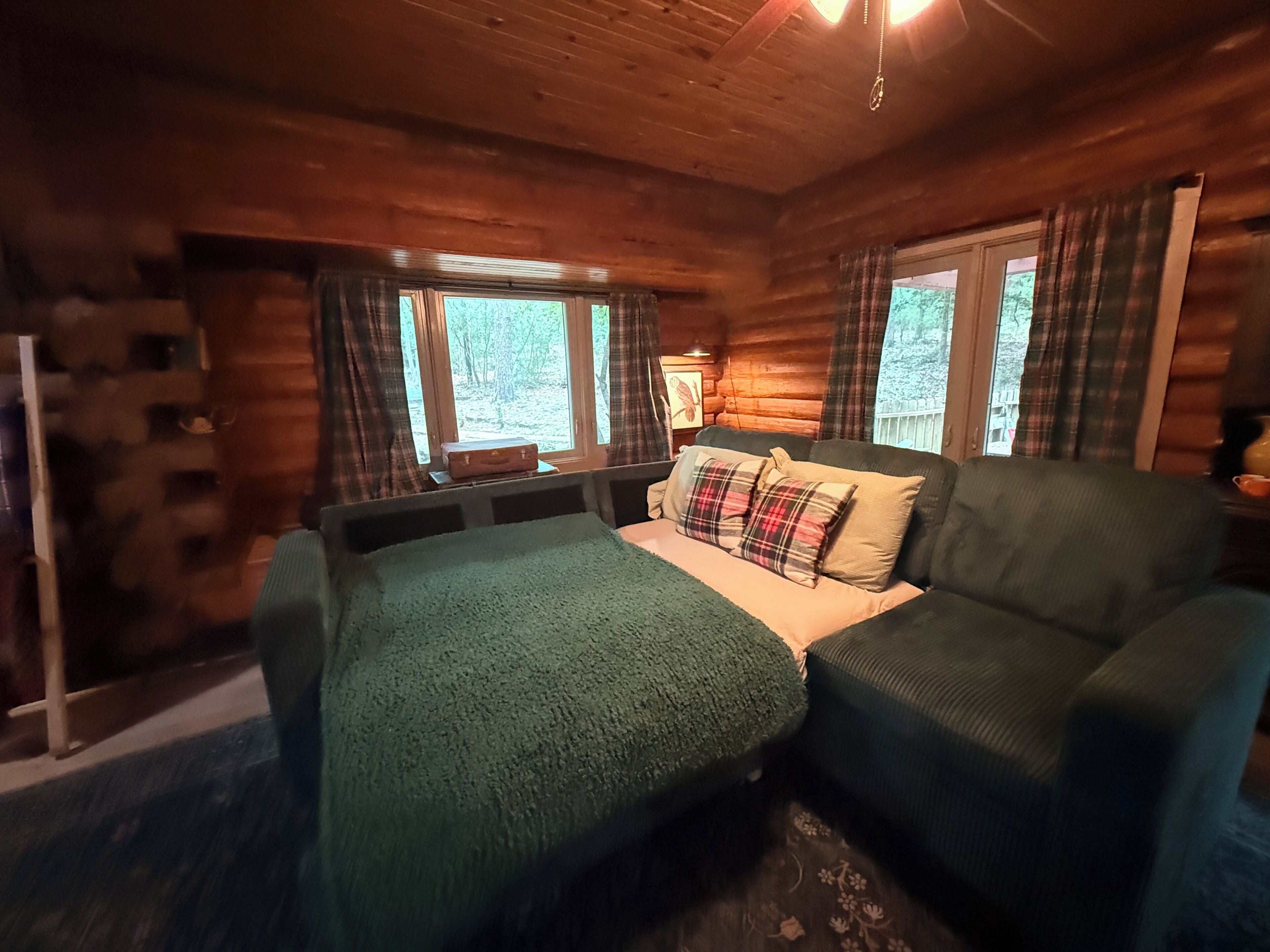 A log cabin room with a bed, a sofa, plaid curtains, and a window with a view of trees.