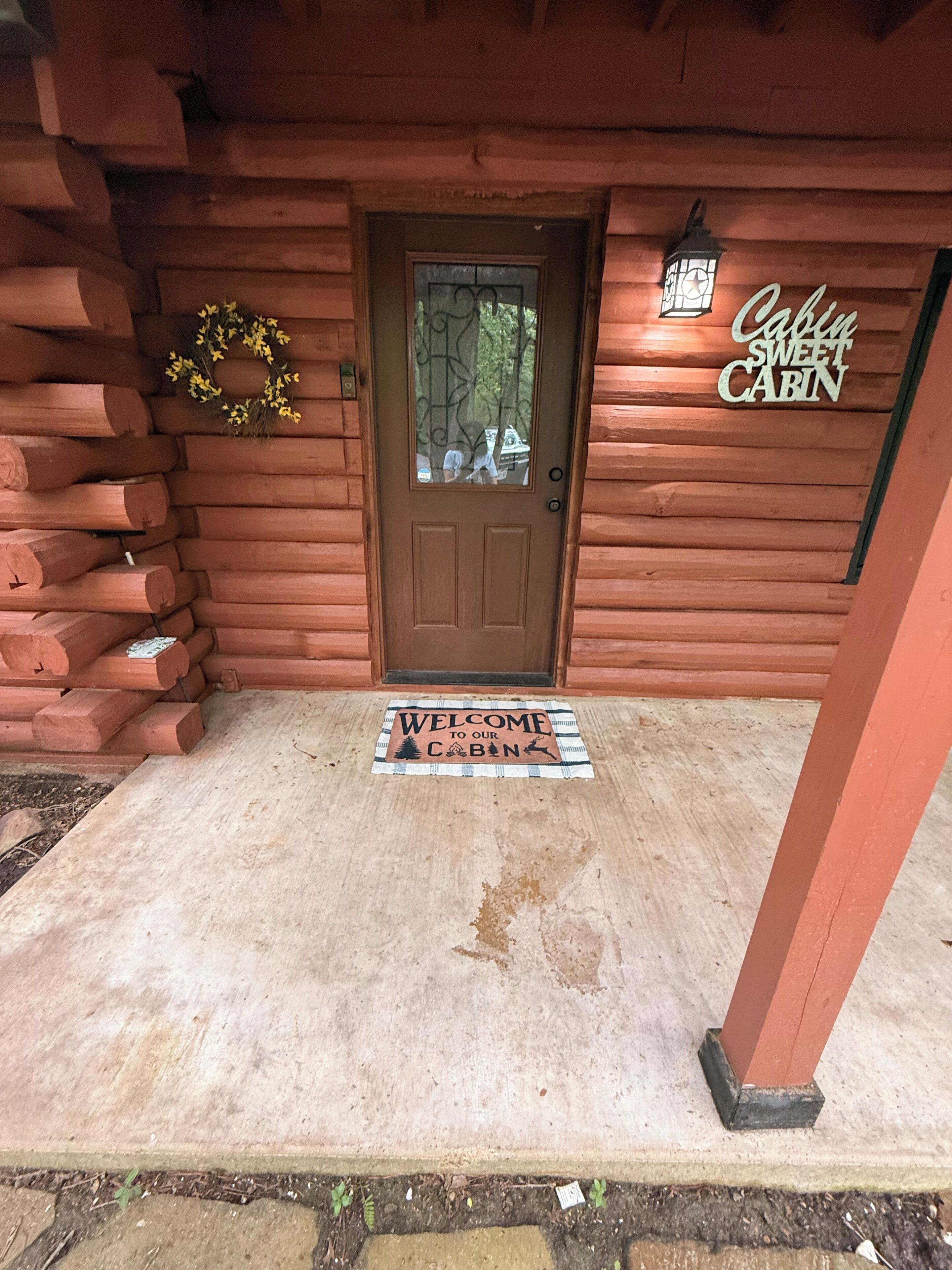 A wooden cabin entrance with a door, a welcome mat, and a sign reading 'Cabin Sweet Cabin.'