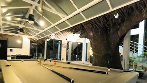 A pool table with a large tree in the background, a modern interior with a high ceiling, and a large window. - Oceana surf Maldives - Kudahuvadhoo (Kudahuvadhoo,)
