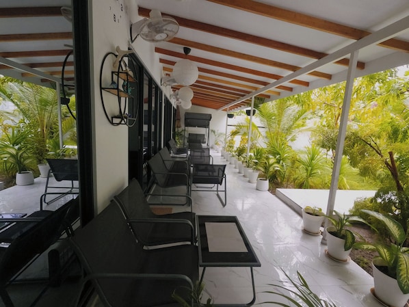 A covered outdoor seating area with tables and chairs, surrounded by potted plants and a view of greenery.