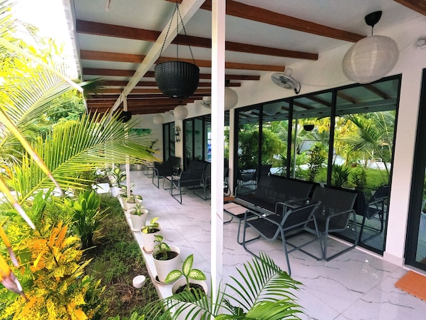 A covered outdoor seating area with chairs and tables, surrounded by potted plants and large windows.