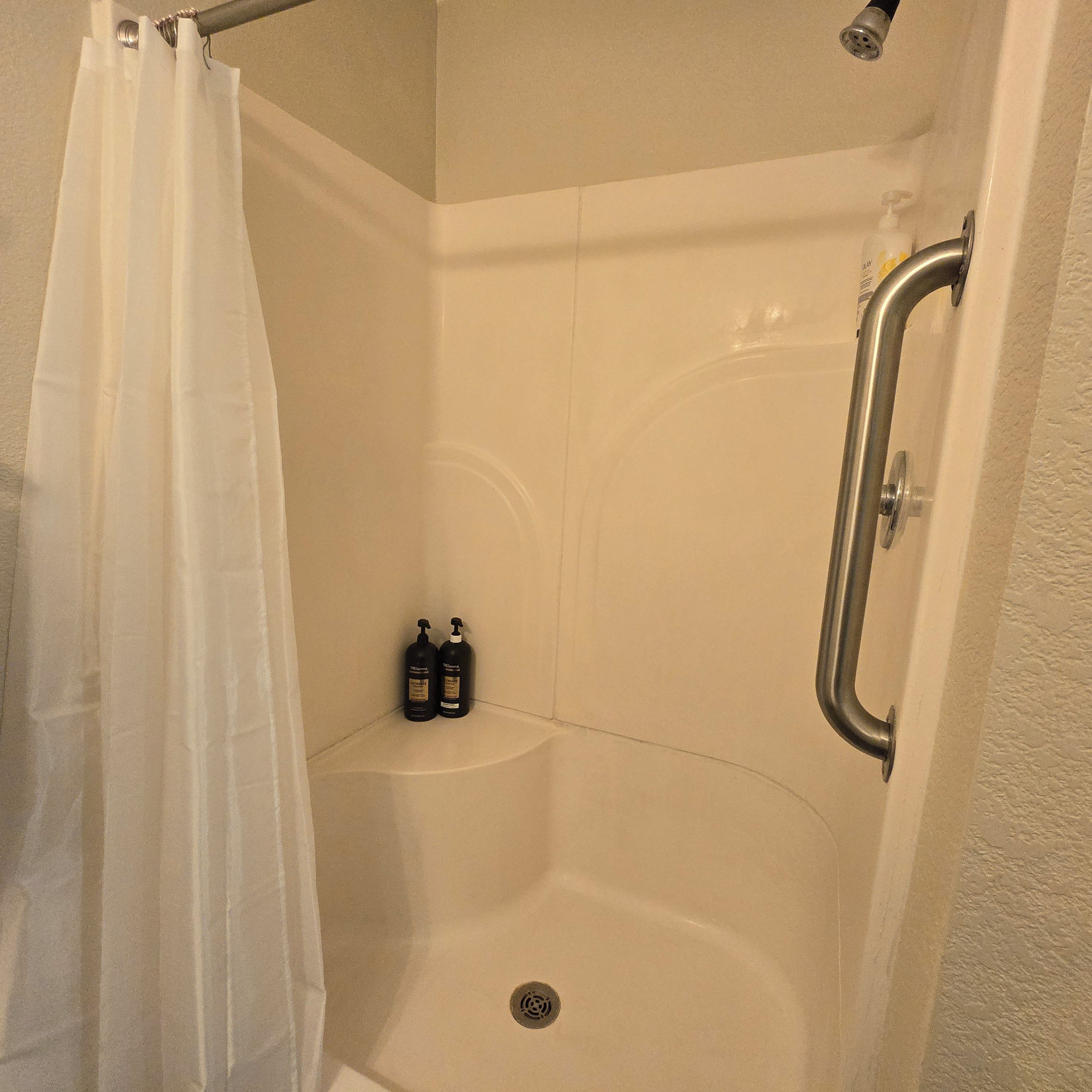 A shower stall with a white curtain, a handheld showerhead, and two bottles of shampoo.