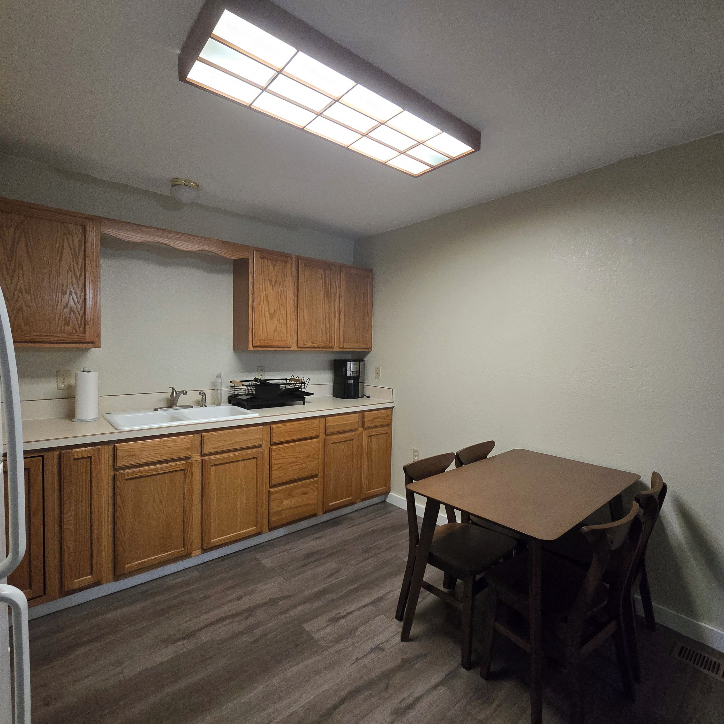 A compact kitchen with wooden cabinets, a white countertop, a coffee maker, and a dining table with chairs.