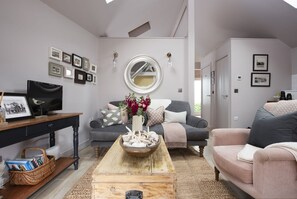Ein Wohnzimmer mit grauem Sofa, einem Holztisch und einem grauen Sessel. An den Wänden hängen Bilder in Rahmen, ein Spiegel und ein kleiner Tisch mit einem Korb.
