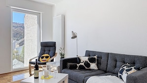 Ein Wohnzimmer mit grauem Sofa, einem Sessel, einem Tisch mit Flasche und Gläsern, einer Stehlampe und Blick durch eine Tür.