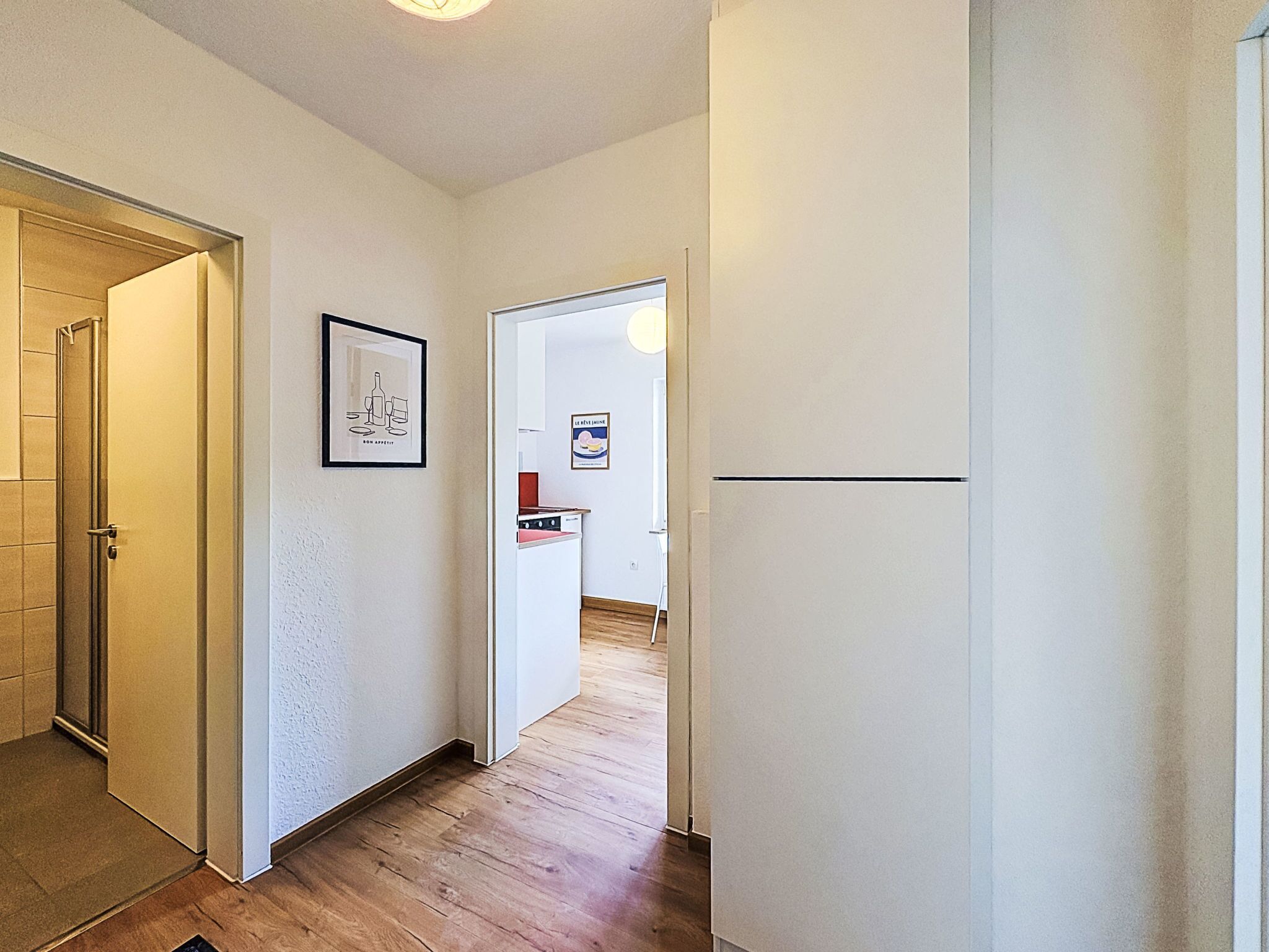 A hallway with a wooden floor, a door on the left, and a refrigerator on the right.