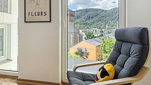 Ein Zimmer mit einem großen Fenster, einem gerahmten Blumenbild und einem grauen Sessel mit Kissen.