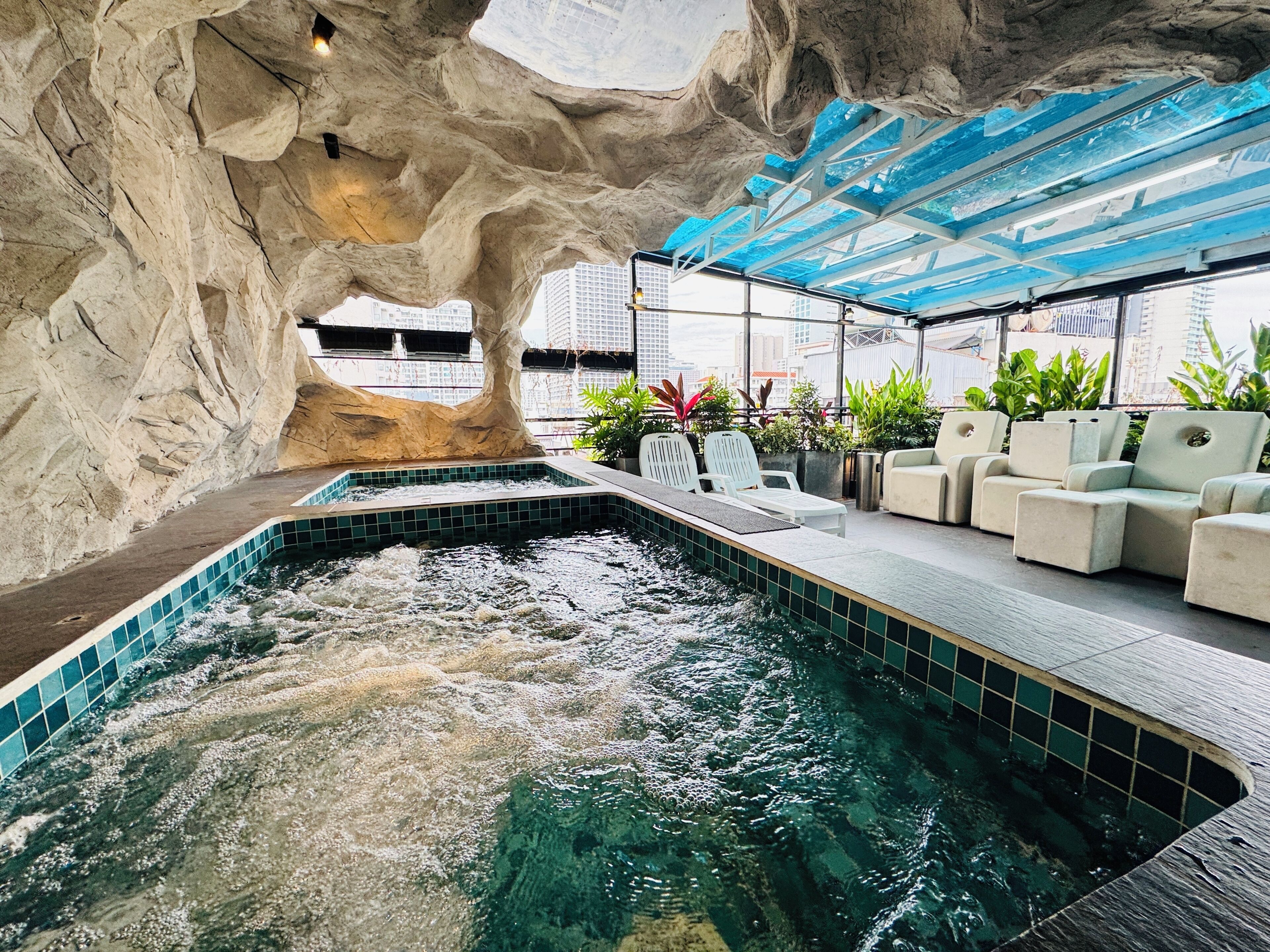 An indoor pool with a rocky cave-like ceiling, lounge chairs, and a view of the cityscape.