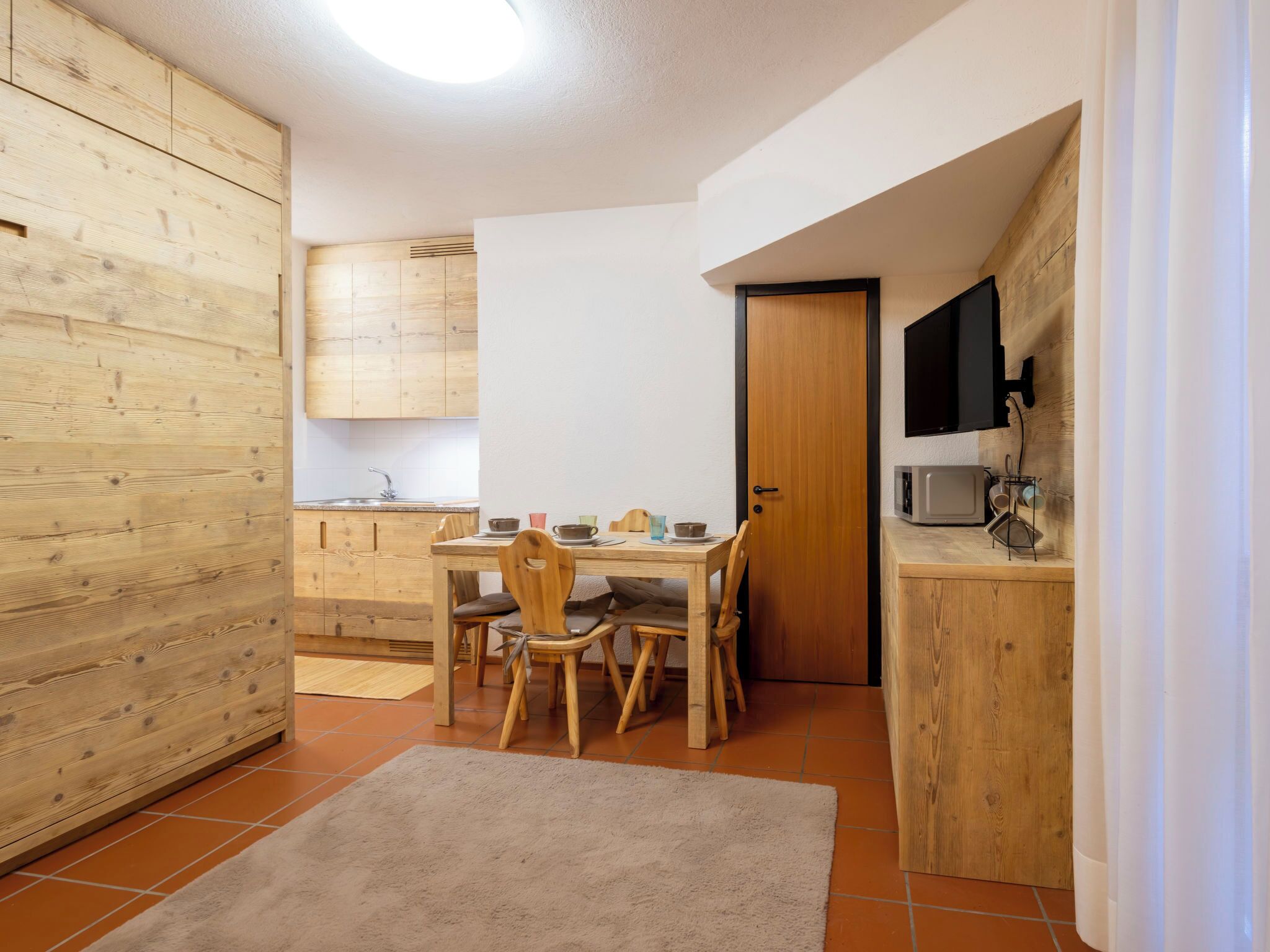 A compact kitchen with wooden cabinets, a dining table with chairs, a TV mounted on the wall, and a microwave on a wooden counter.