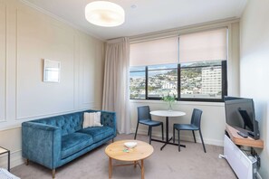 Een woonkamer met een blauwe bank, een ronde houten tafel, twee stoelen, een televisie en een raam met uitzicht op de stad.