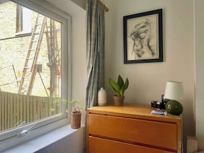 Ein Zimmer mit Fenster, einem Holzkasten, einem gerahmten Kunstwerk, einer Topfpflanze und einer Lampe.