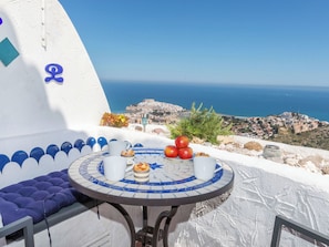 Ein runder Tisch mit einer blauen und weißen Mosaikplatte, eine blaue gepolsterte Bank und ein malerischer Blick auf das Meer sowie eine Stadt im Hintergrund.