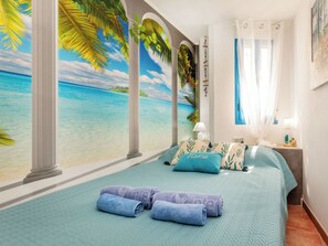 Ein Schlafzimmer mit großem Fenster, einem Bett mit blauer Bettwäsche und einem tropischen Wandgemälde.