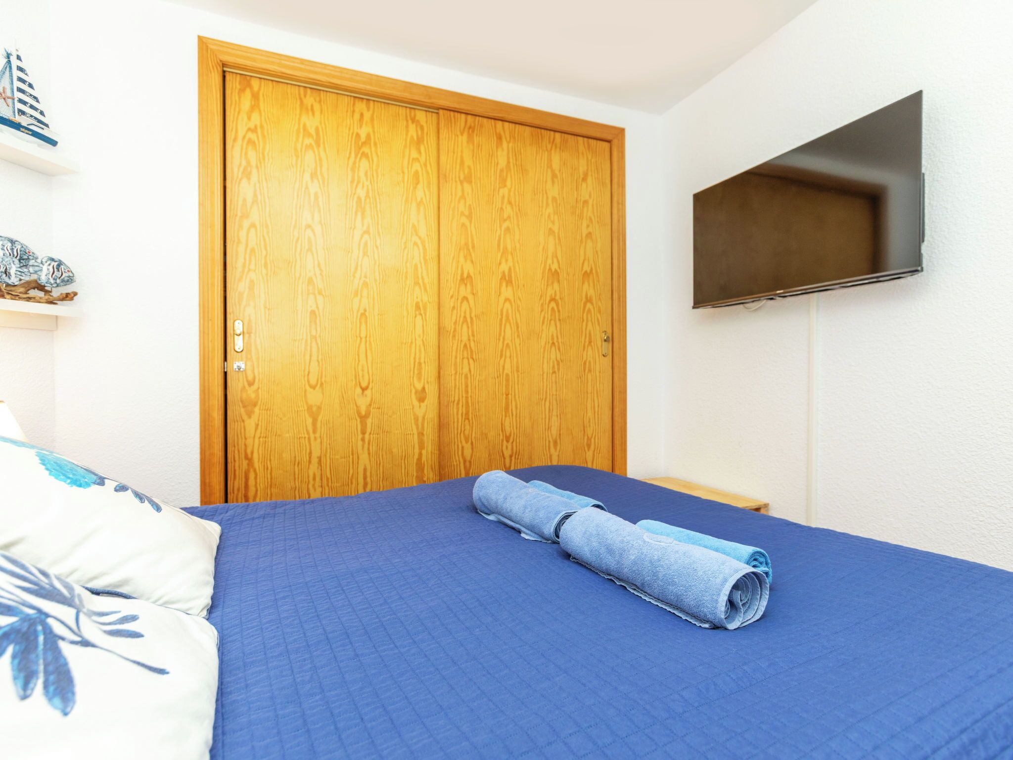 A bedroom with a wooden door, a bed with a blue bedspread, and a flat-screen TV mounted on the wall.