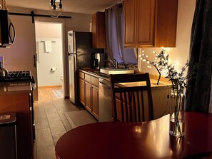 A compact kitchen with wooden cabinets, a refrigerator, a coffee maker, and a dining table with a vase of flowers.
