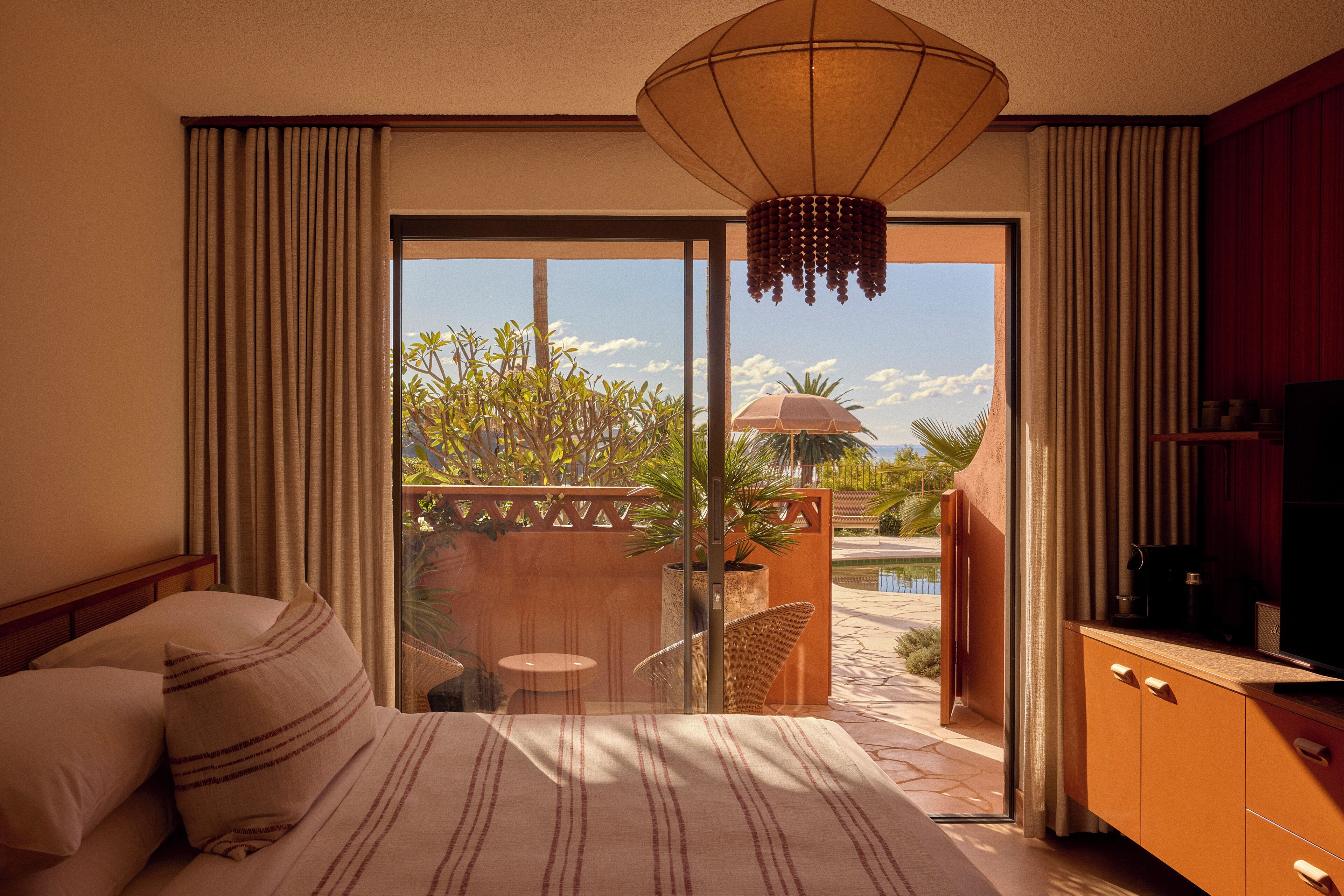 Camera d'albergo con un letto, una TV e un balcone vista piscina e vegetazione.