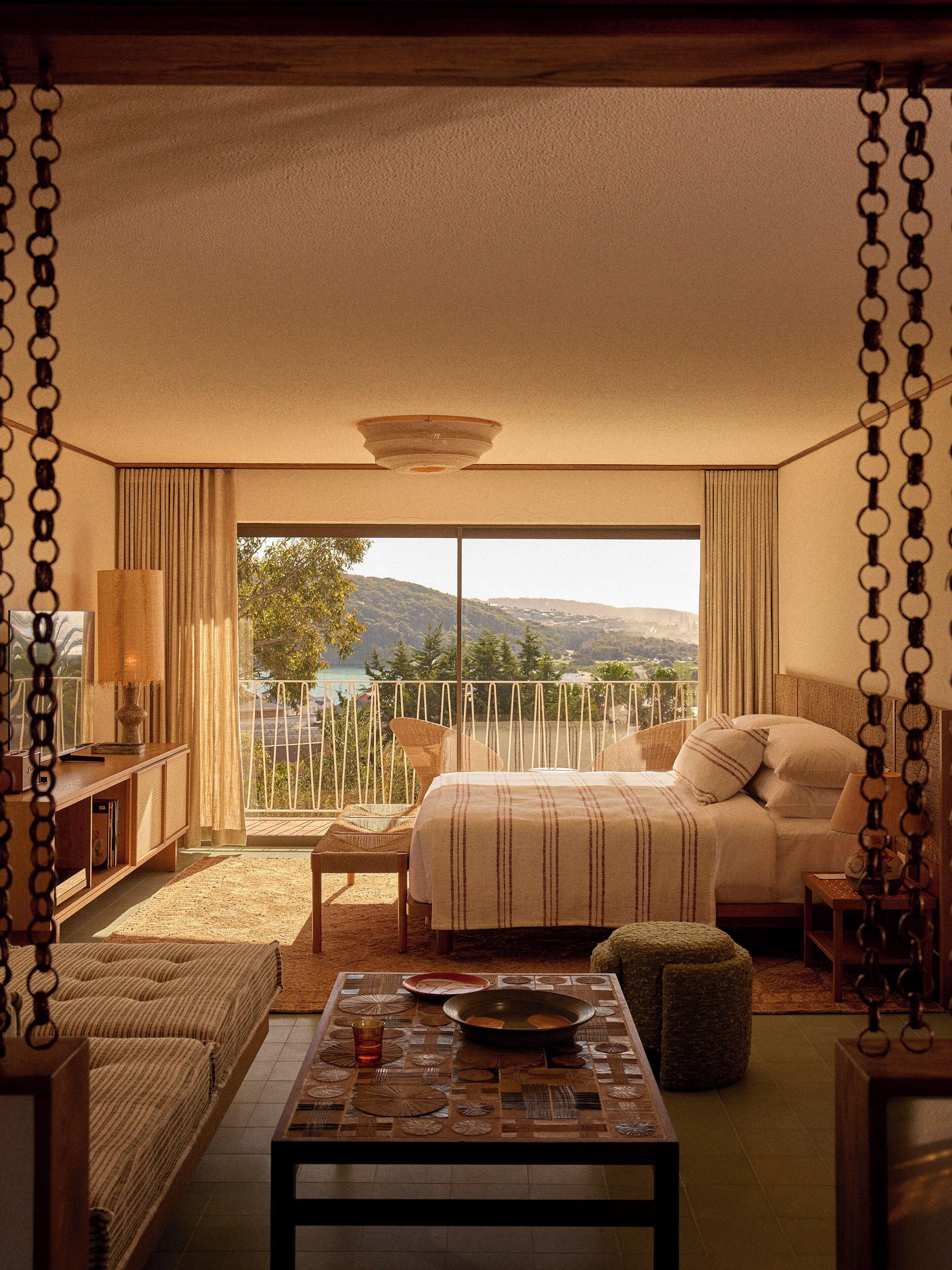 Quarto com cama, mesa de cabeceira, cadeira e vista para o exterior.