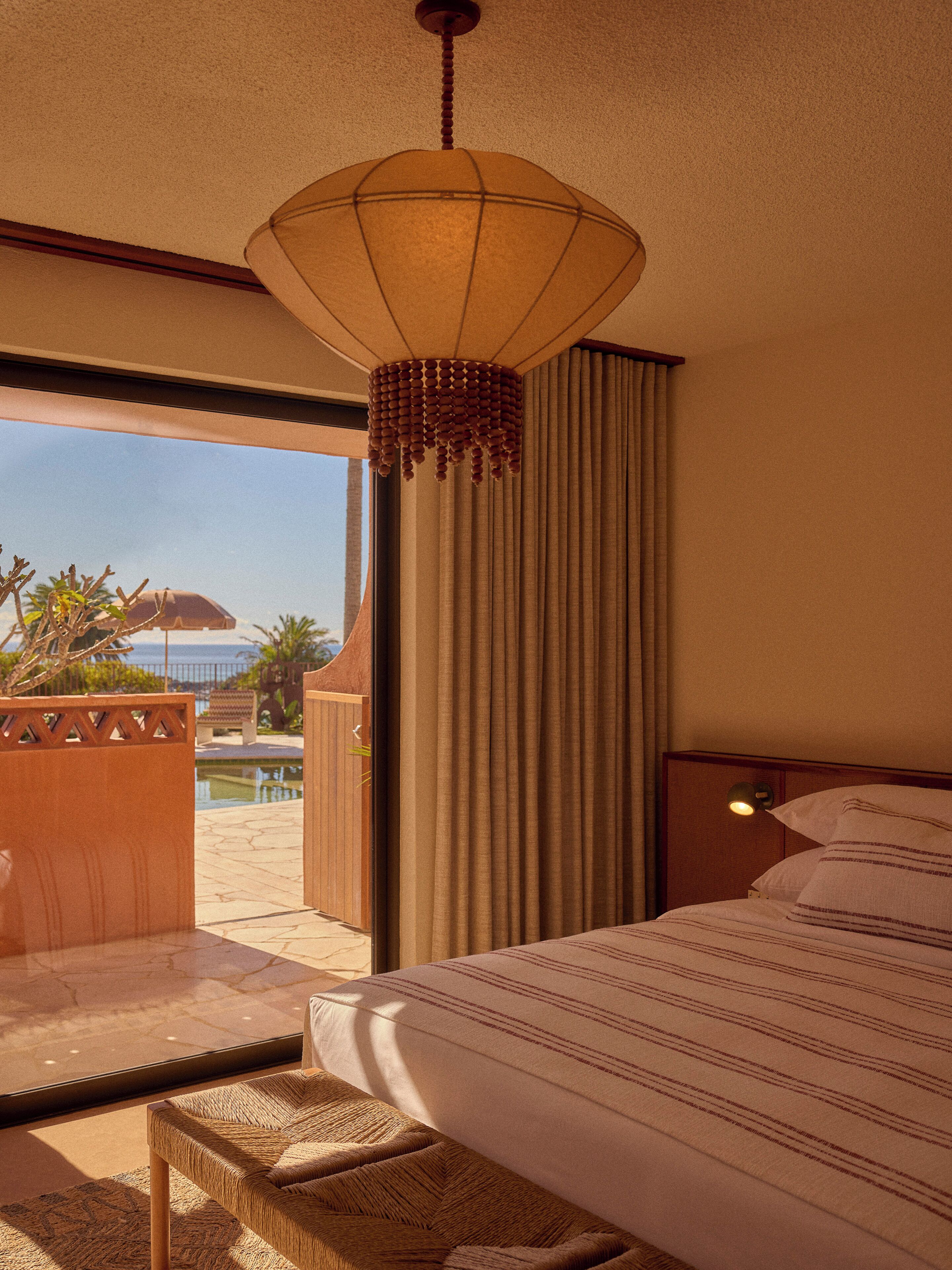 Una habitación de hotel con una cama, una lámpara colgante, vistas a una piscina y un banco de madera.