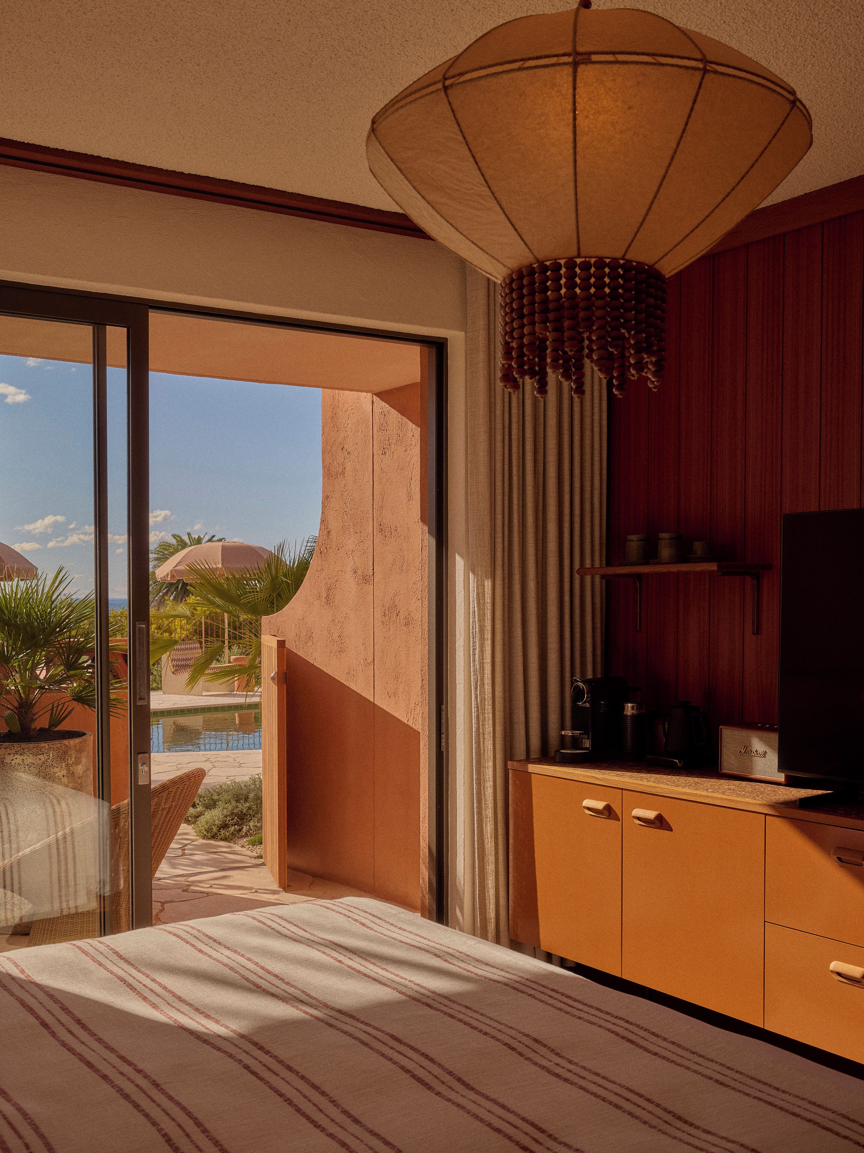 A hotel room with a bed, a television, a minibar, and a view of a pool and palm trees.