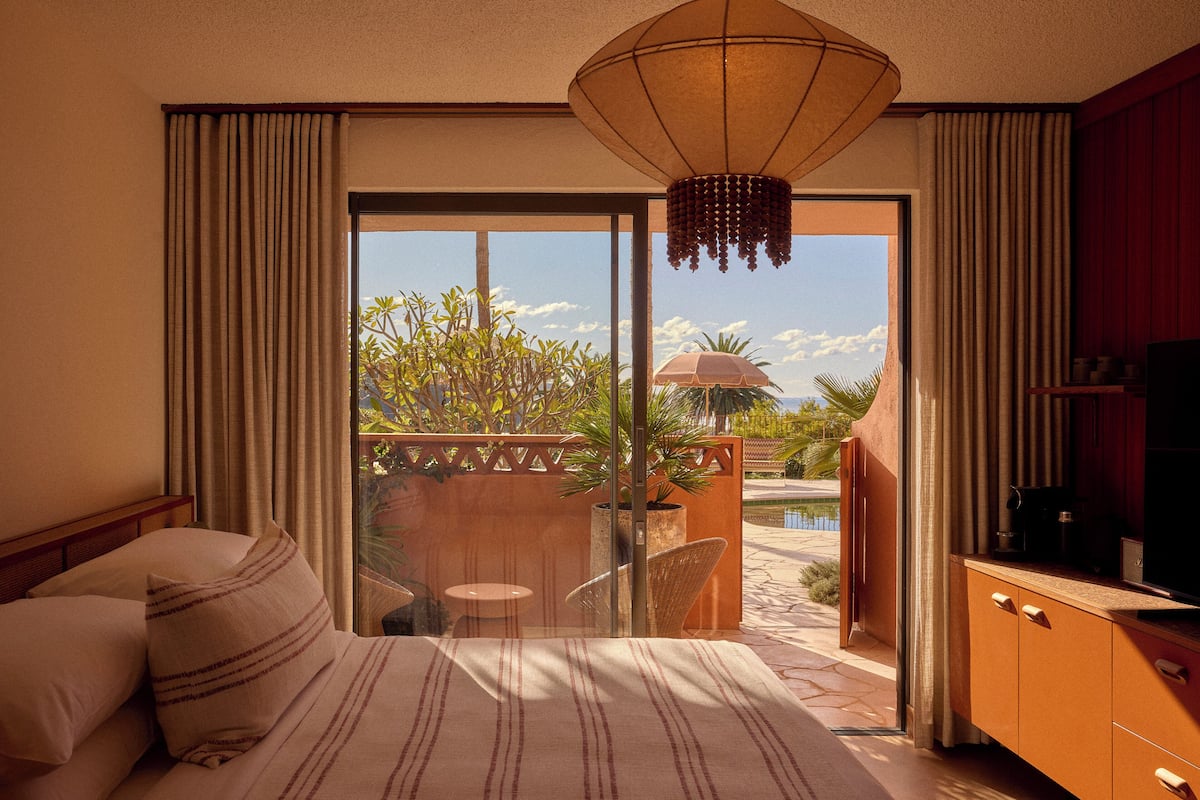 A hotel room with a bed, a TV, a balcony with a view of the pool and greenery.